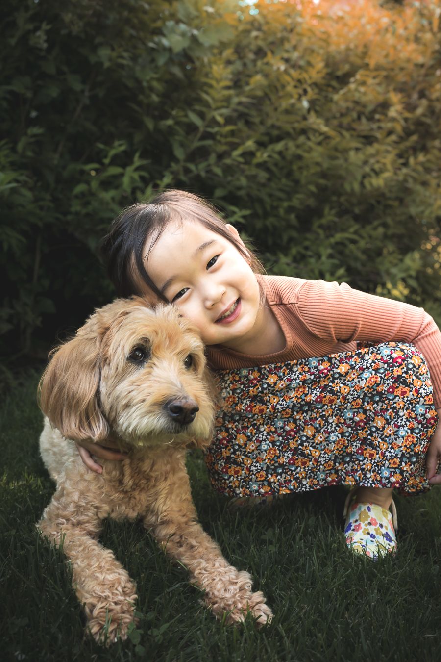Smiling young Asian girl hugging a fluffy golden doodle dog on green grass with lush bushes in background.