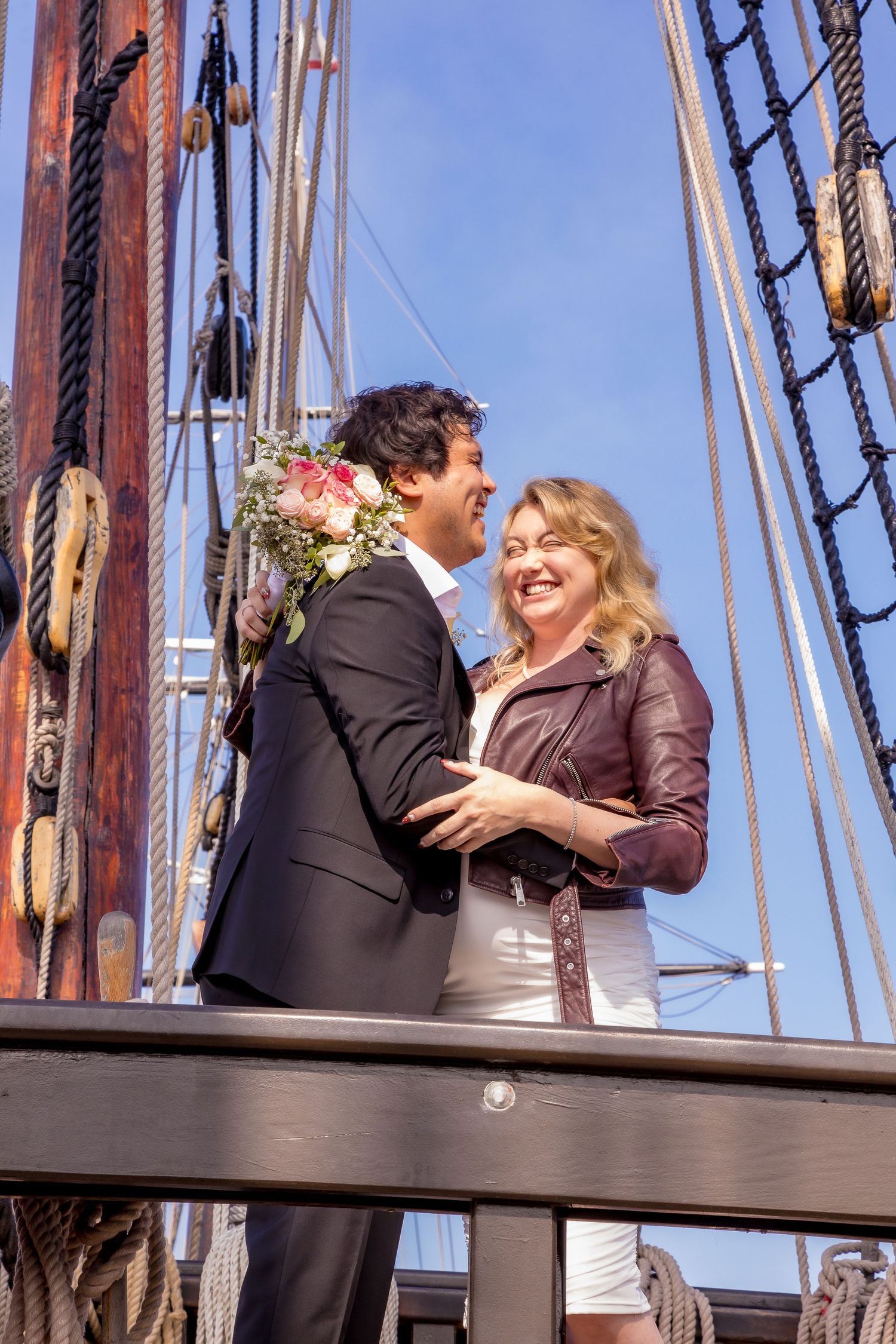 Happy couple embracing on a tall ship deck, man holding flowers, woman in leather jacket, blue sky background.