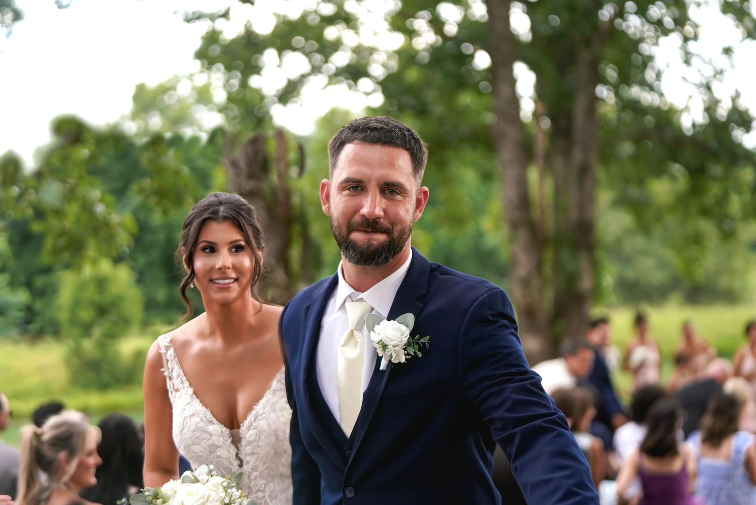 Bride and groom smiling outdoors at wedding ceremony, groom in navy suit, bride in lace gown, green trees behind.