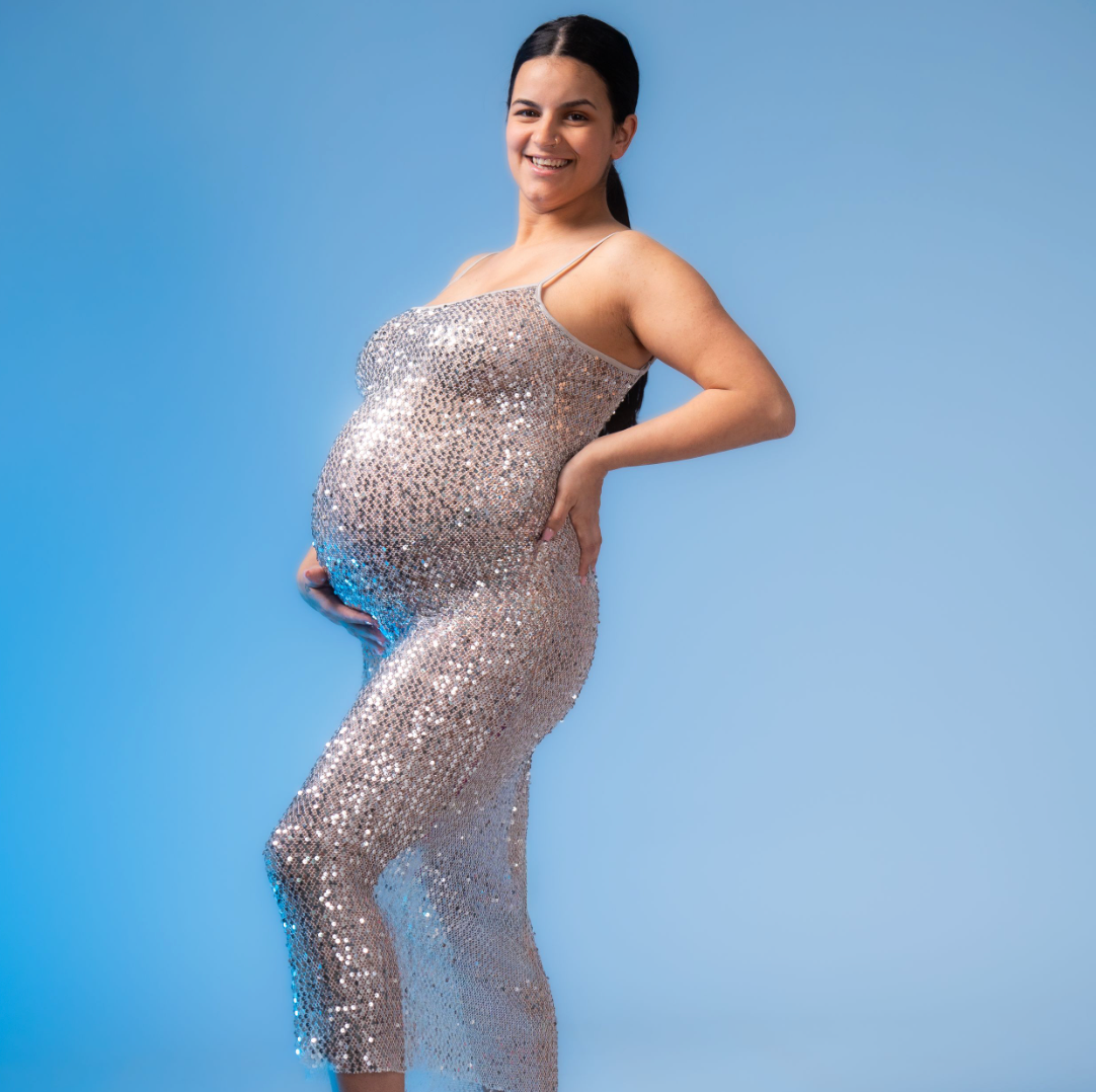 Pregnant woman in a sparkling sequin maternity gown poses gracefully against a blue background.