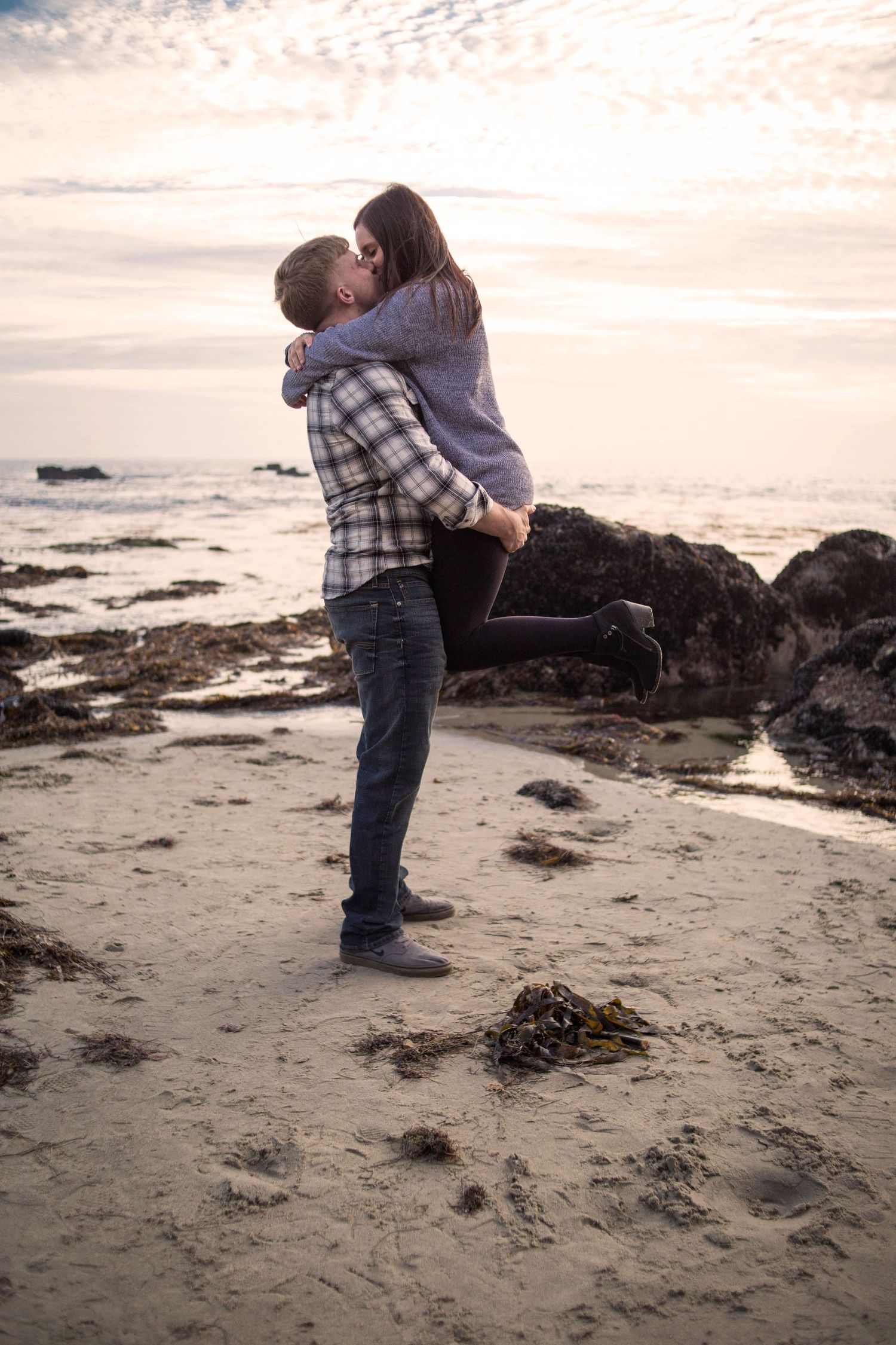 Couple embracing and kissing on a rocky beach at sunset during an engagement photo session.