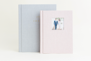Two elegant linen wedding photo albums in soft blue and blush pink, one featuring a couple portrait window on the cover.