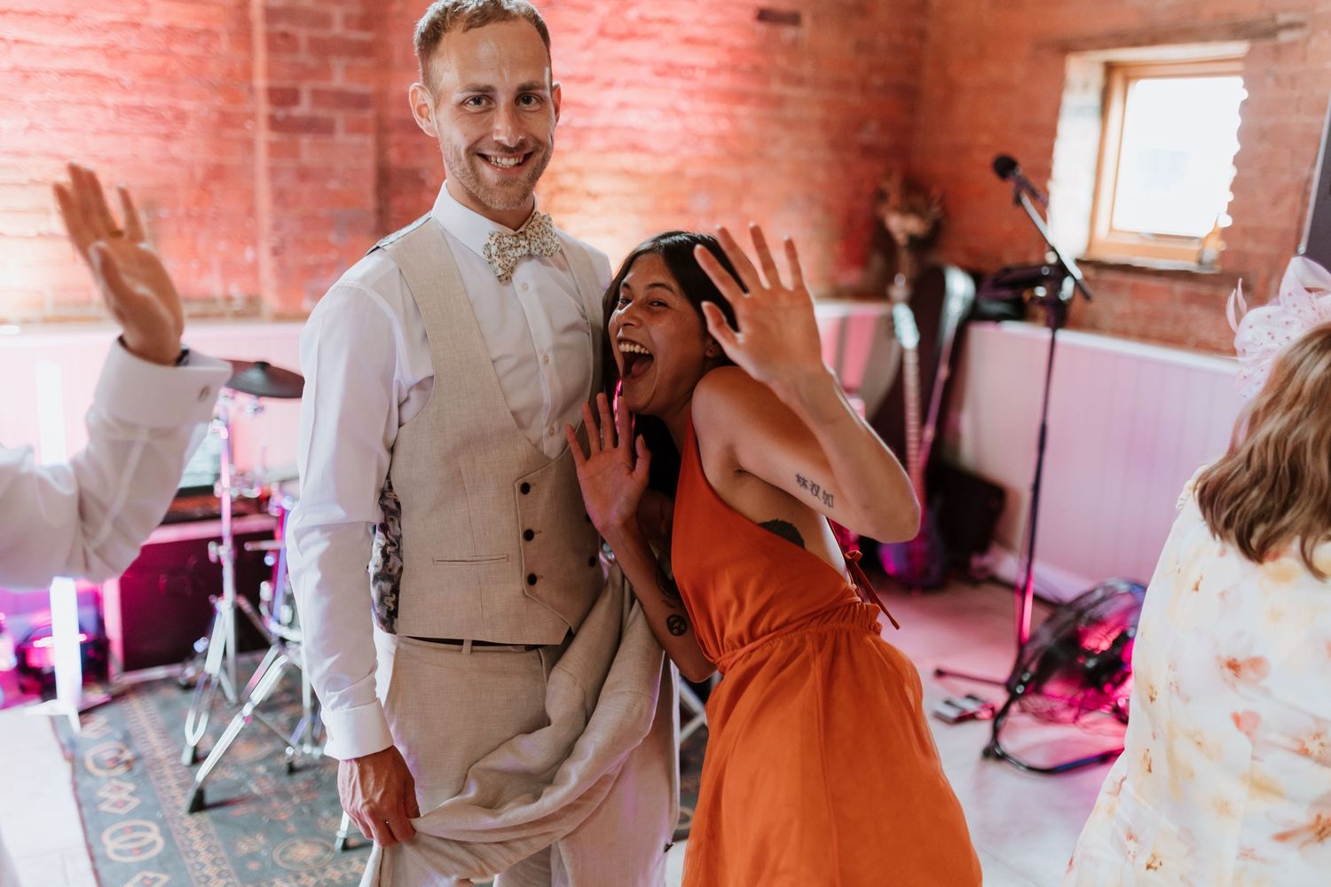 Groom and friends dancing joyfully at a wedding reception in a rustic brick venue, woman in orange dress, man in gray vest.
