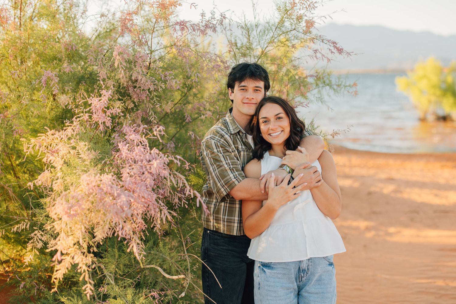 Couple sitting together on red sand dunes with a lake and mountains in the background on a sunny day.