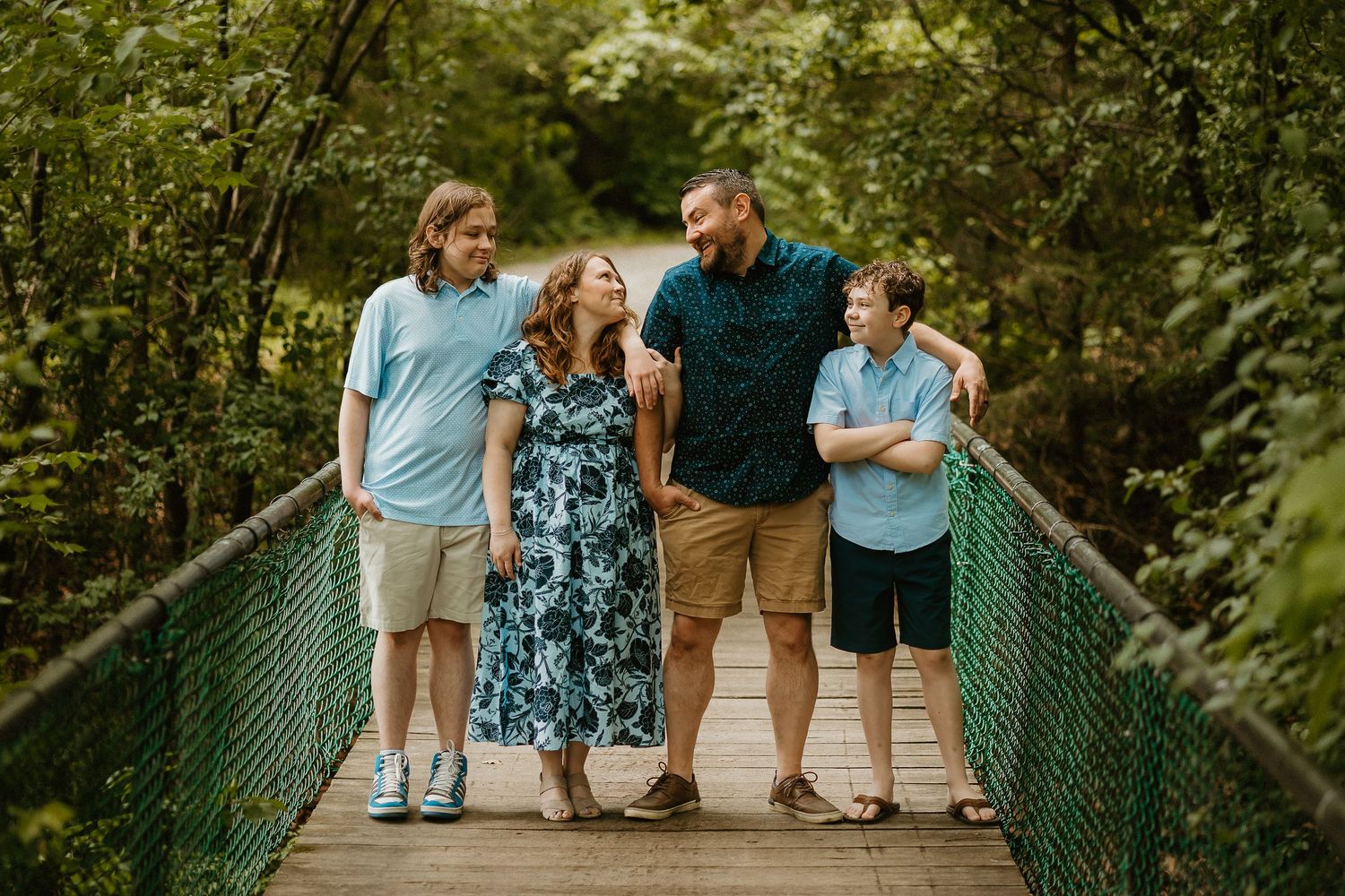 Family of four smiling on a wooden bridge surrounded by lush green trees during an outdoor photo session.