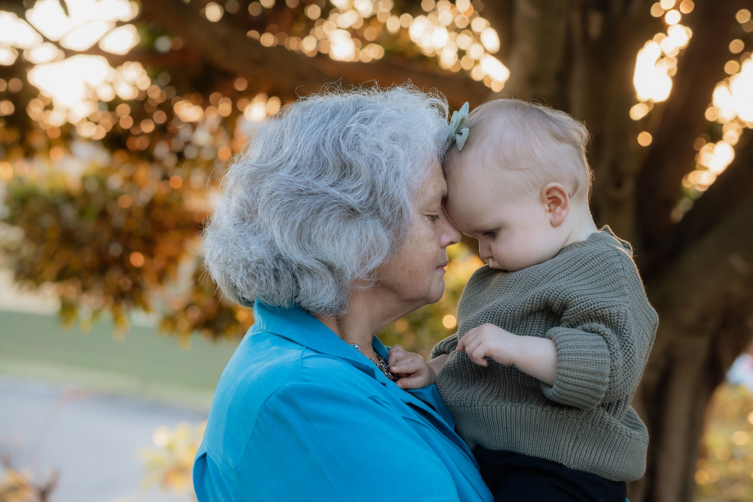 Elderly grandmother with gray hair tenderly holds and nuzzles a baby outdoors in warm golden light photoshoot in Beaumont Hills.