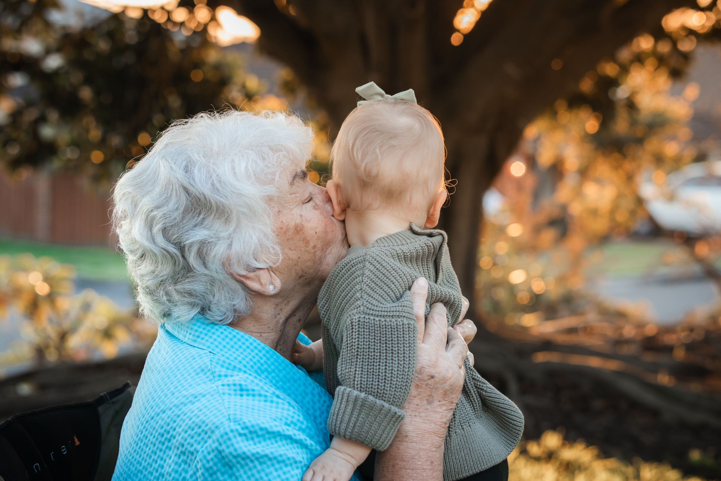 Elderly grandmother tenderly kissing a baby in a green sweater outdoors near a tree in warm golden light at Turkey's nest, Beaumont Hills.