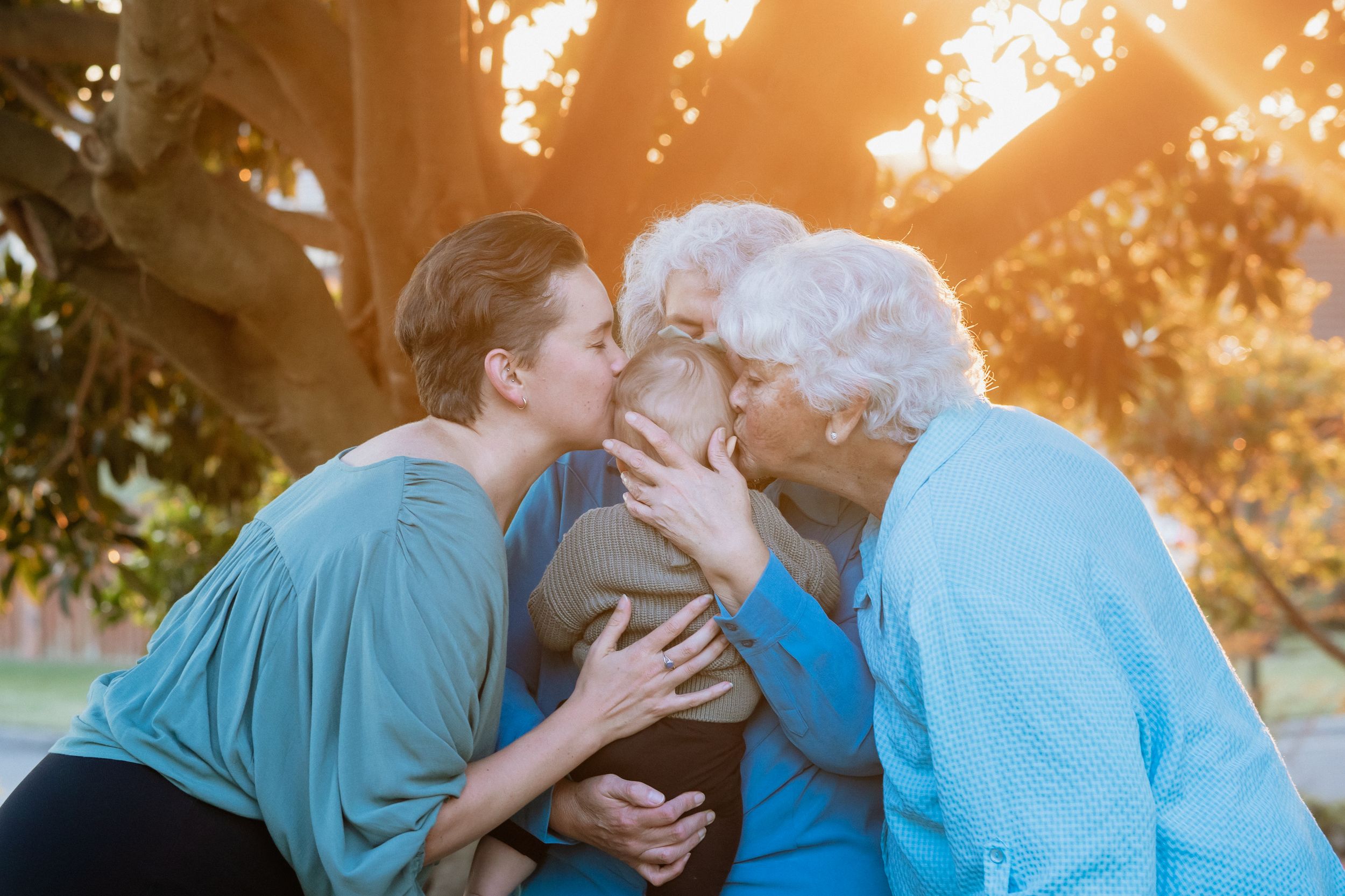 A woman and elderly grandmothers share kisses on a baby outdoors under a glowing tree at sunset in Turkey's Nest Reserve, Beaumont Hills.