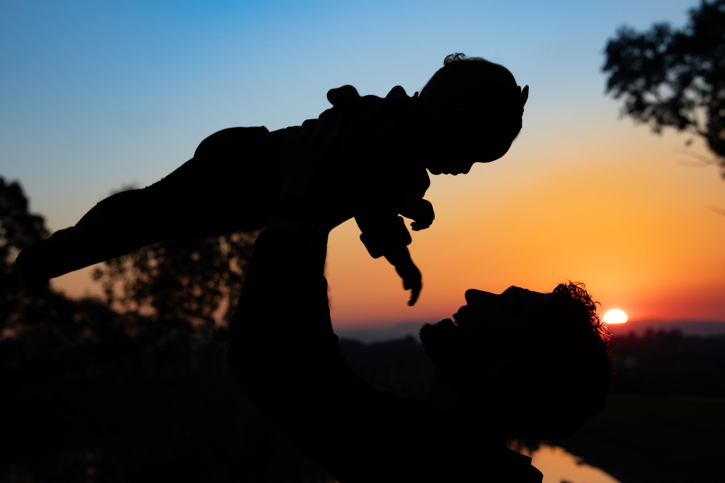 Silhouette of parent lifting baby against vibrant sunset sky with orange and blue hues in Beaumont Hills.