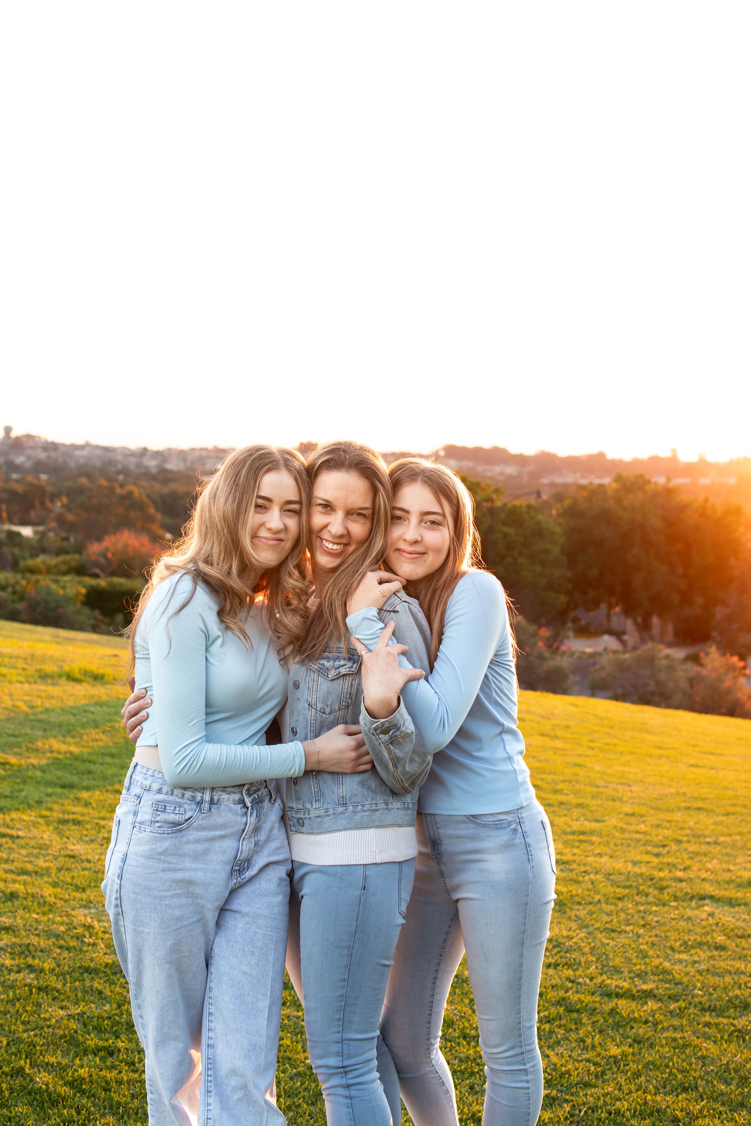 Two teenage girls in blue jeans and tops hugging their mum outdoors at golden hour sunset on green grass overlooking Rouse Hill.