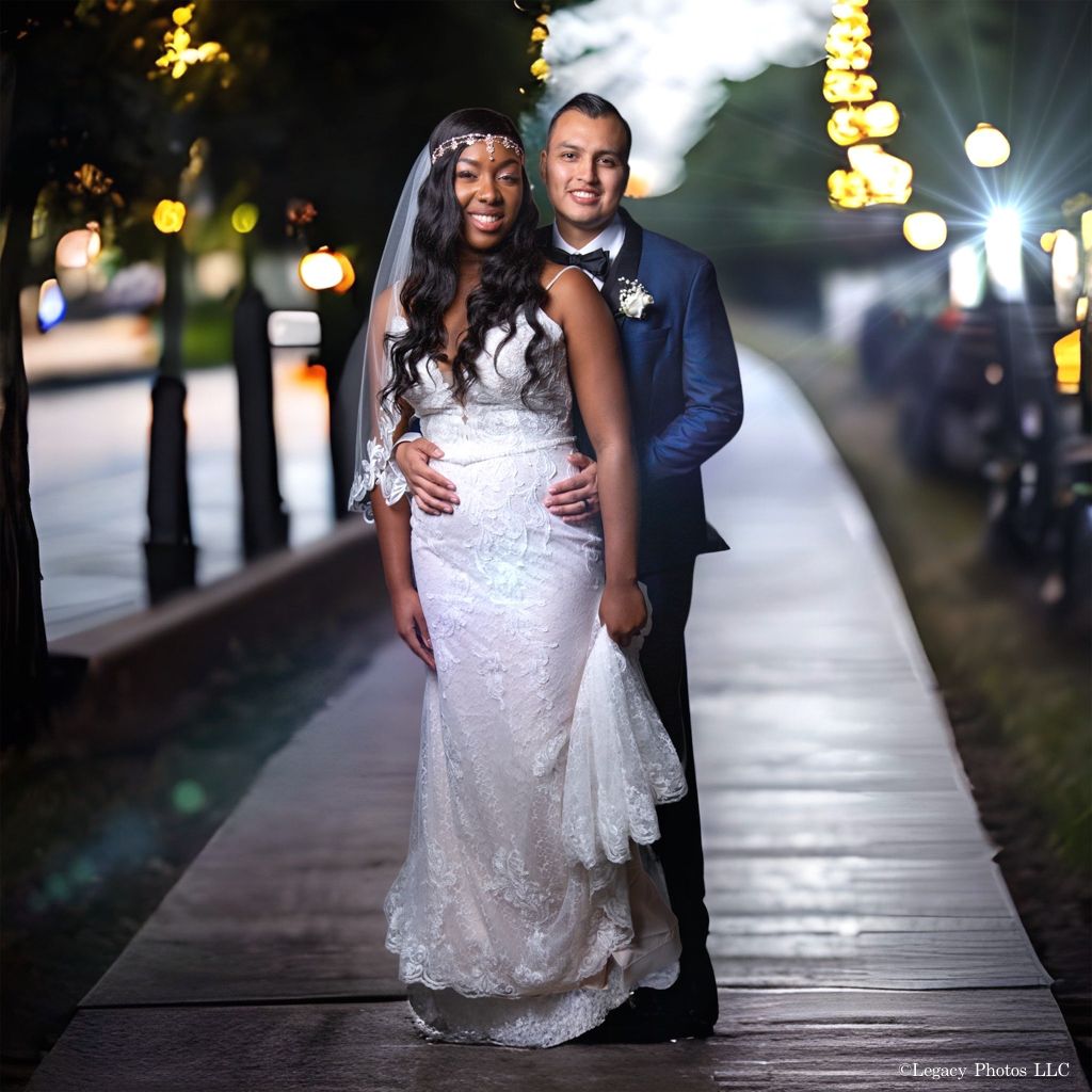 Bride and groom standing together on a tree-lined walkway with warm lights behind them.
