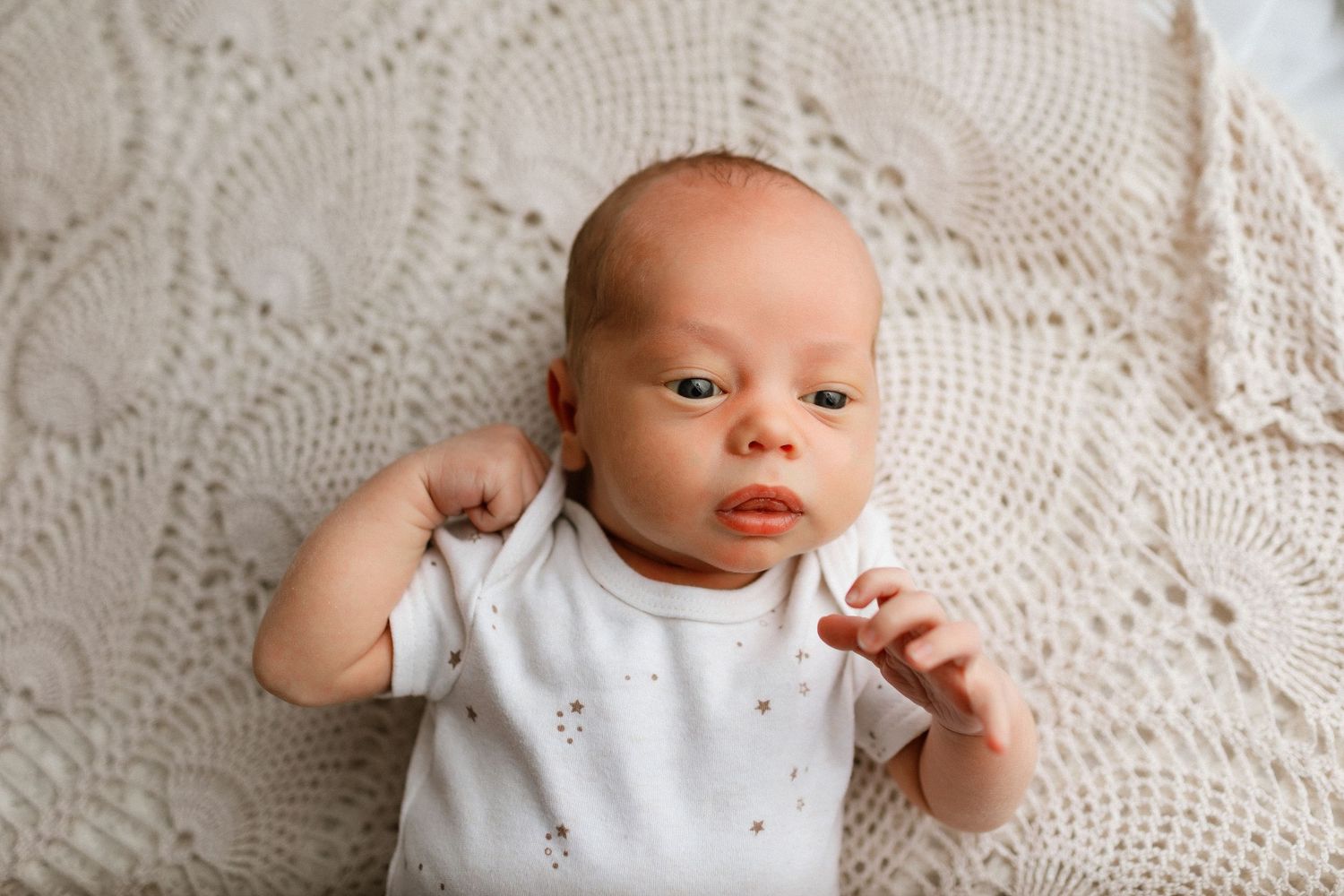 Newborn baby in white onesie lying on cream crochet blanket, looking alert with tiny fist raised.