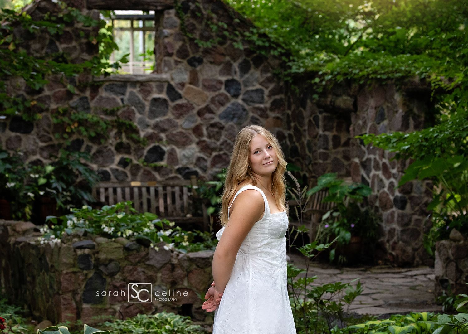 Young blonde girl in white dress poses in lush garden with stone wall backdrop, professional portrait photography.