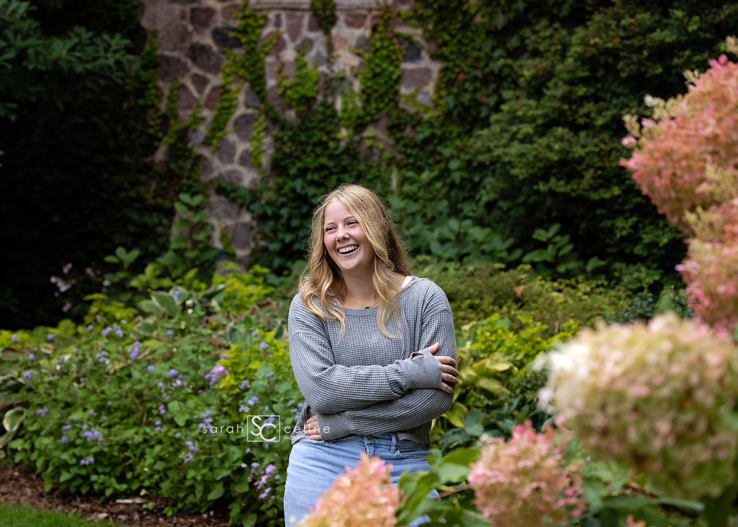 Laughing teenage girl in gray sweater poses with arms crossed in a lush garden with pink hydrangeas.