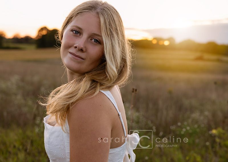 Young blonde woman in white dress posing in golden hour sunlit field for senior portrait photography.