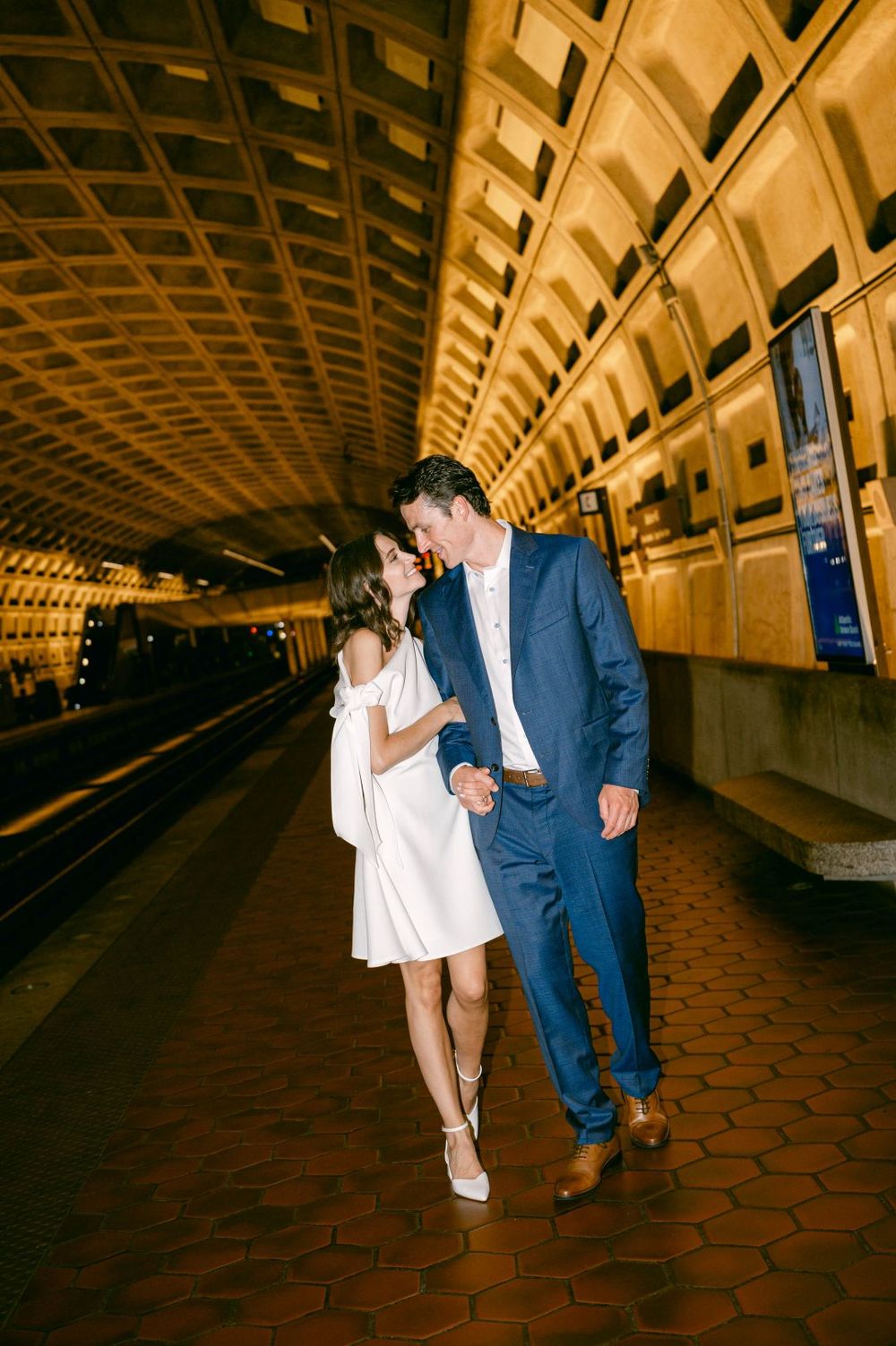 Couple in white dress and blue suit holding hands and smiling at each other on a DC Metro subway platform.
