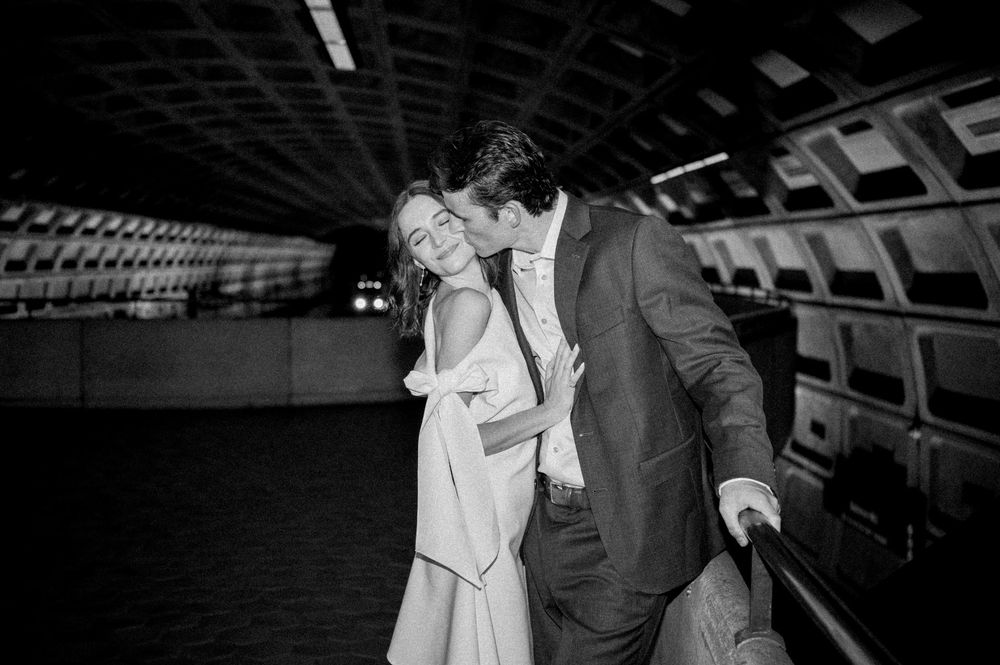 Black and white photo of a couple embracing and kissing in a metro station, dressed in formal wedding attire.