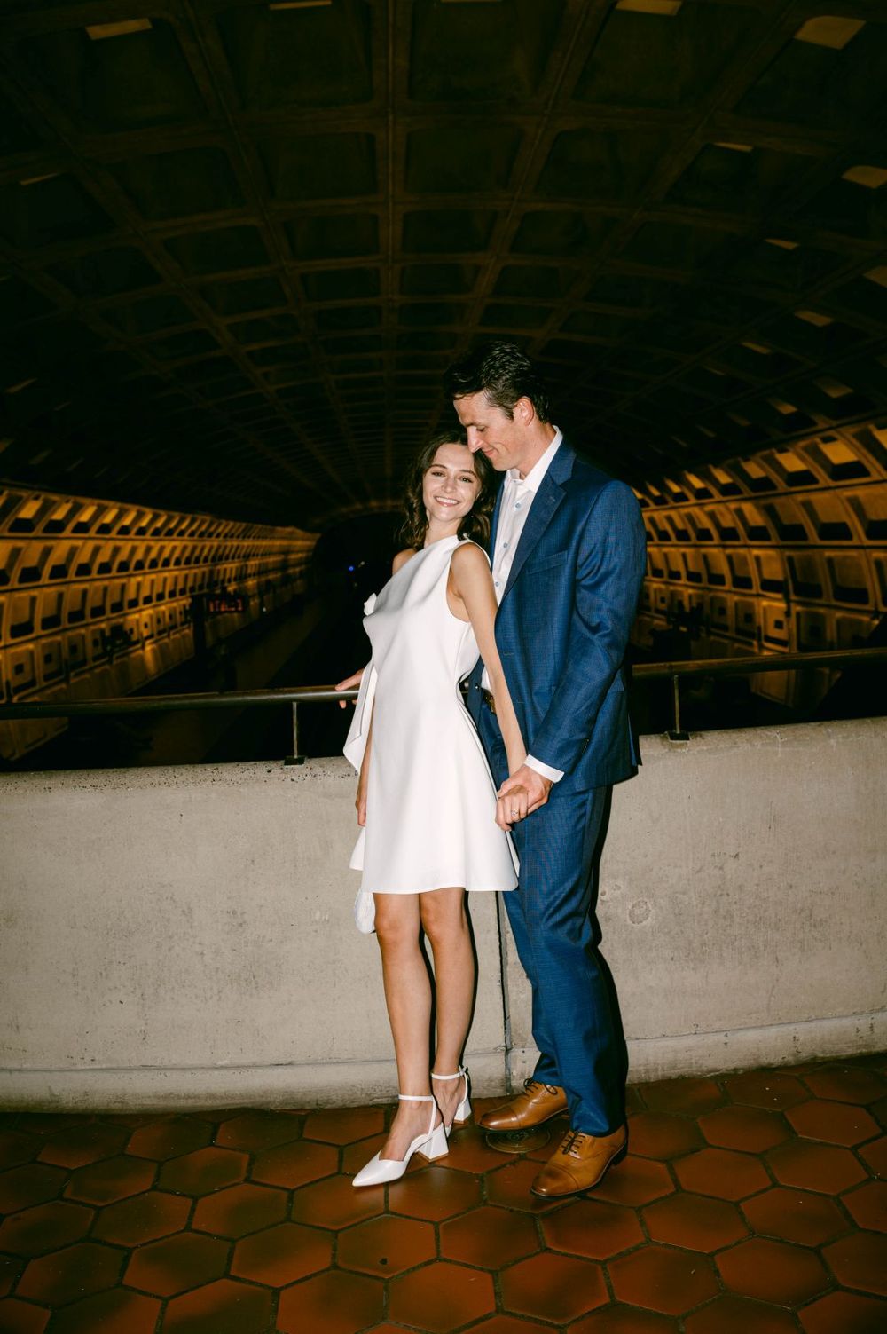 Couple in wedding attire posing in a DC Metro station, woman in white dress and man in navy suit holding hands.