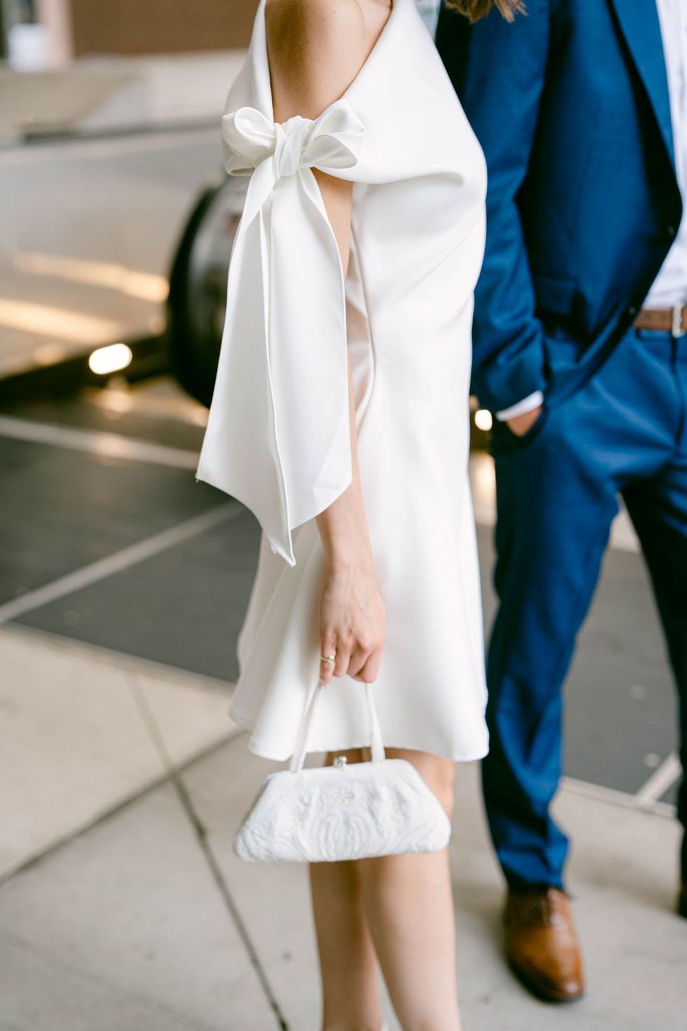 Bride in white mini dress with bow back detail holds beaded clutch beside groom in navy suit.