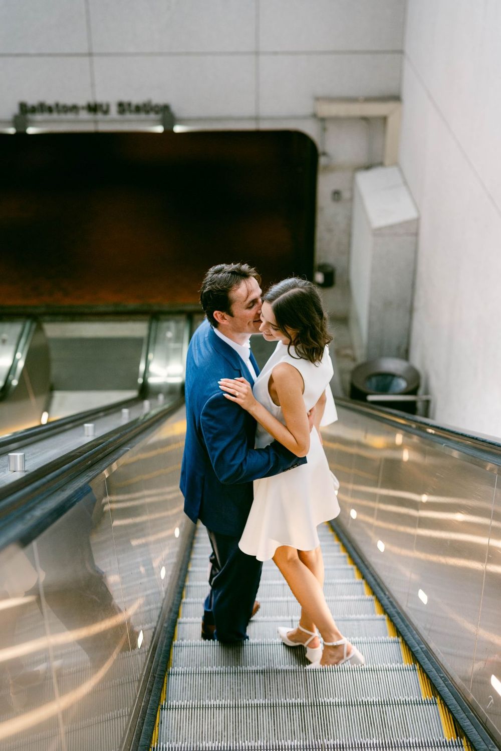 Couple sharing a romantic moment on an escalator at Ballston-MU Metro Station, man in blue suit, woman in white dress.