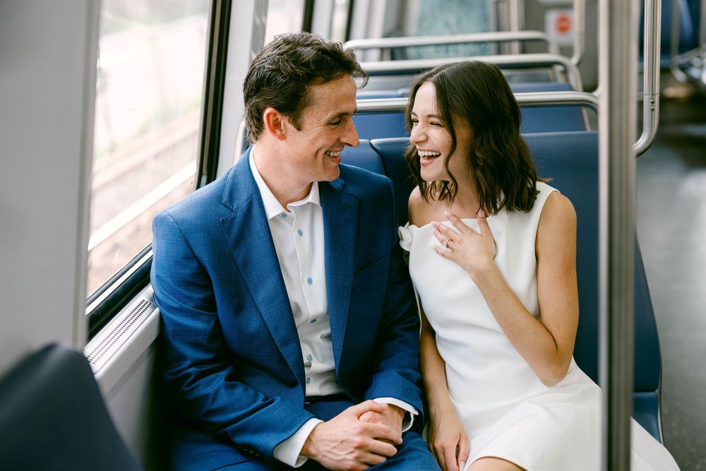 Couple laughing together on a subway train, man in blue suit and woman in white dress, sharing a joyful moment.
