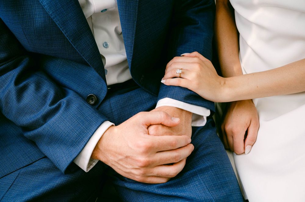Bride wearing diamond ring holds groom's hand in blue suit, close-up wedding detail photo.