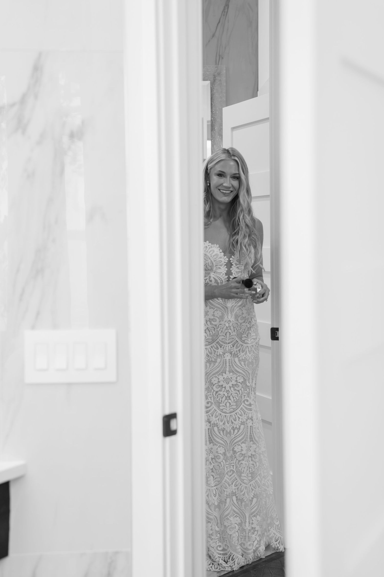 Black and white photo of a smiling bride in a lace wedding gown peeking through a doorway.