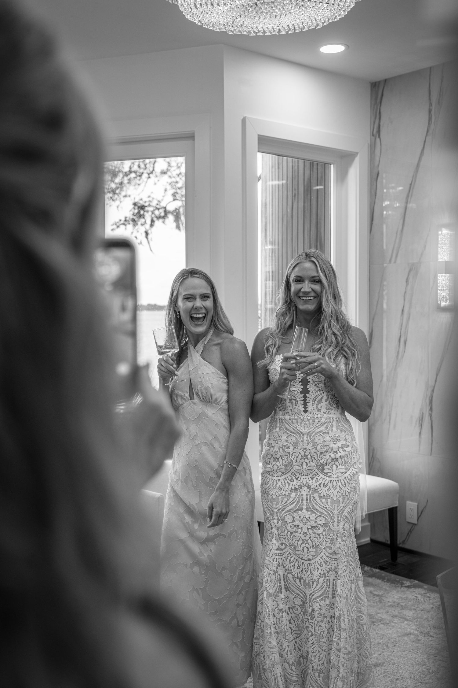 Two bridesmaids in lace gowns laugh joyfully holding champagne glasses during a wedding getting-ready moment.