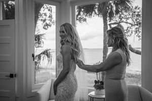 Black and white photo of bride in lace wedding dress being helped by bridesmaid near window with palm trees.