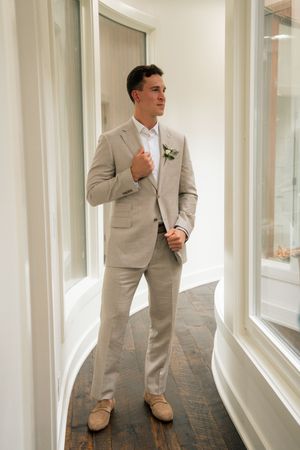 A groom in a beige linen suit with a white boutonniere stands by large windows on dark hardwood floors.