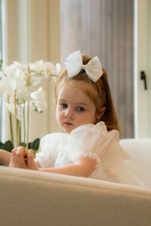 Adorable toddler girl in white dress and bow hair clip sitting near white orchids.