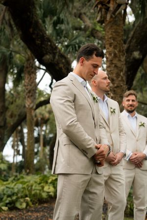 Groom in beige linen suit stands with groomsmen outdoors under trees at garden wedding ceremony.