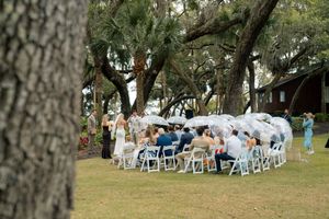Outdoor wedding ceremony under sprawling oak trees with white chairs and guests gathered on a lush green lawn.