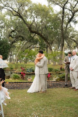 Bride and groom share first kiss at outdoor wedding ceremony under Spanish moss oak trees near a garden pond.