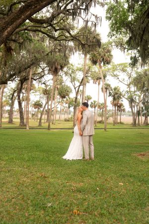 Bride and groom kissing under Spanish moss-draped oak trees with palm trees in background on lush green lawn.