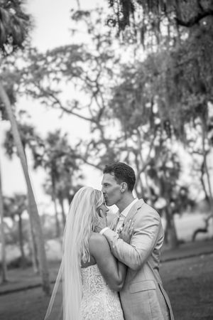Black and white photo of a bride and groom sharing a romantic kiss outdoors among moss-draped trees.