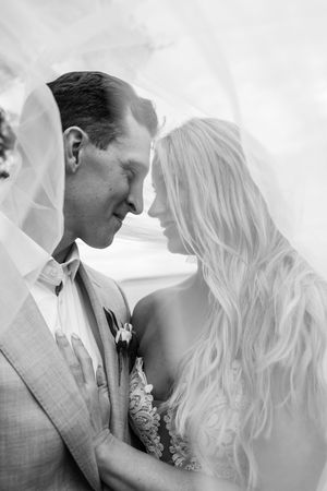 Black and white photo of bride and groom touching foreheads under a flowing veil on their wedding day.