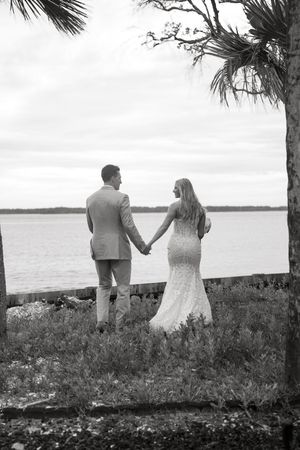 Black and white photo of a bride and groom holding hands facing a serene waterfront, framed by palm trees.