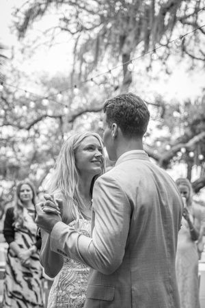 Black and white photo of a bride and groom sharing their first dance outdoors under trees with string lights.