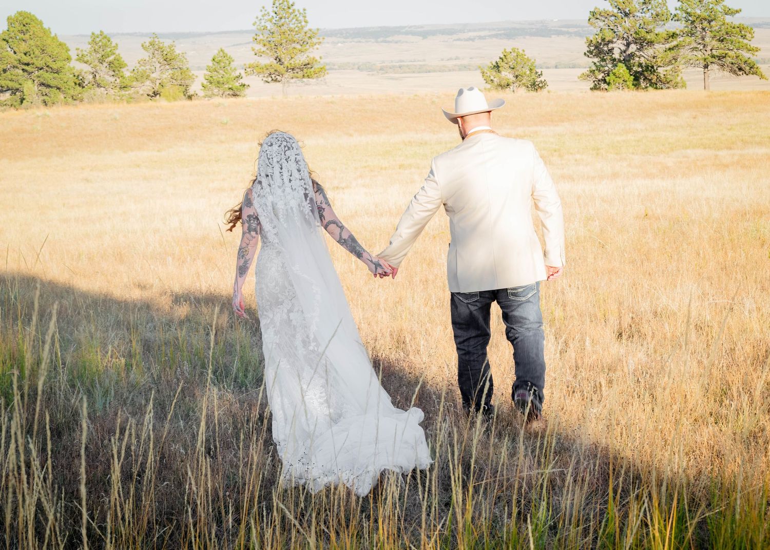Couple in western wedding attire embracing in a field, groom in cowboy hat, bride holding colorful feather bouquet.