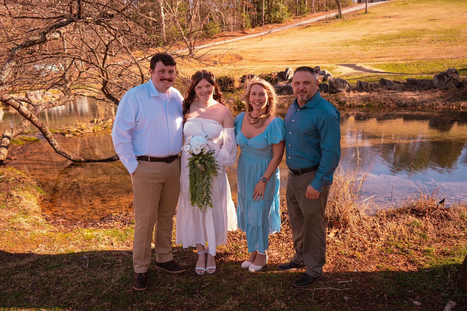 Family of four posing outdoors near a pond, with bride in white wedding dress holding bouquet, on a sunny day.