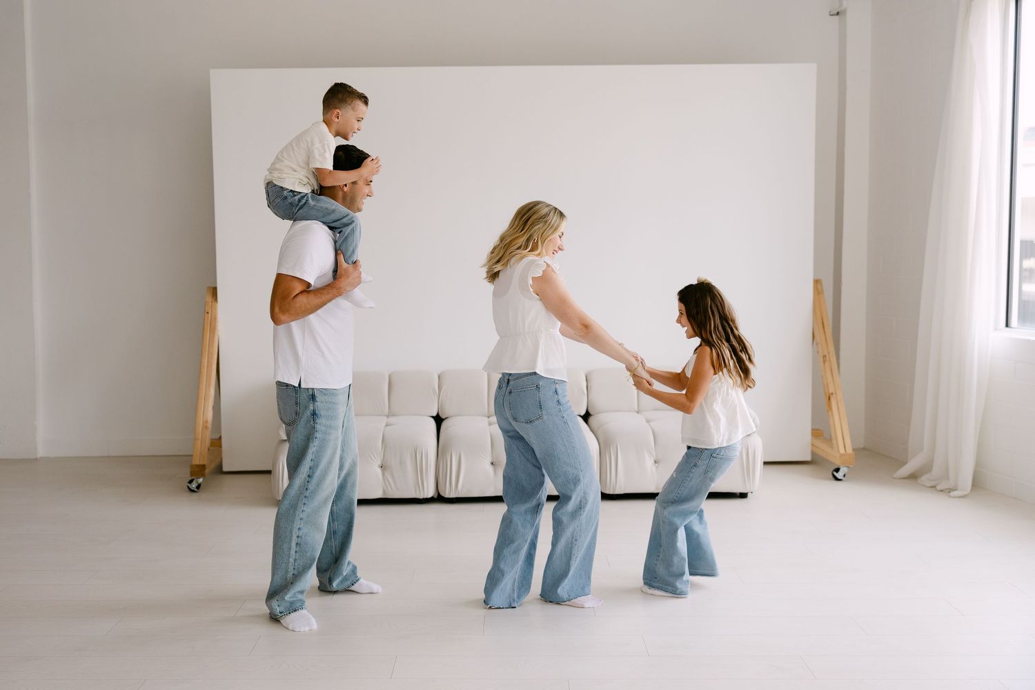 Family of four in white tops and jeans playing together in a bright white studio space with a couch.