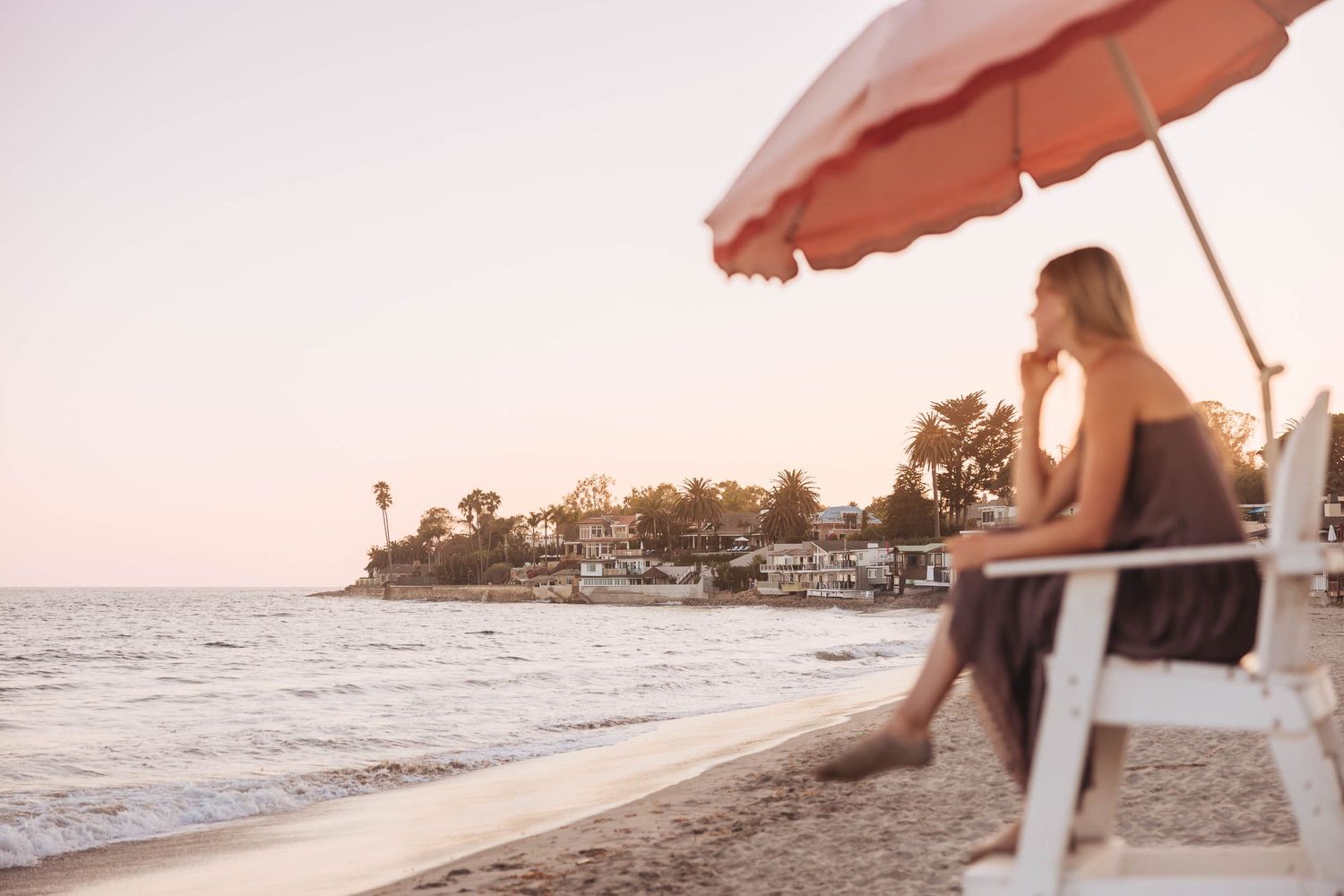 Woman sitting under a red beach umbrella at sunset, gazing at the ocean with a coastal town in the background.