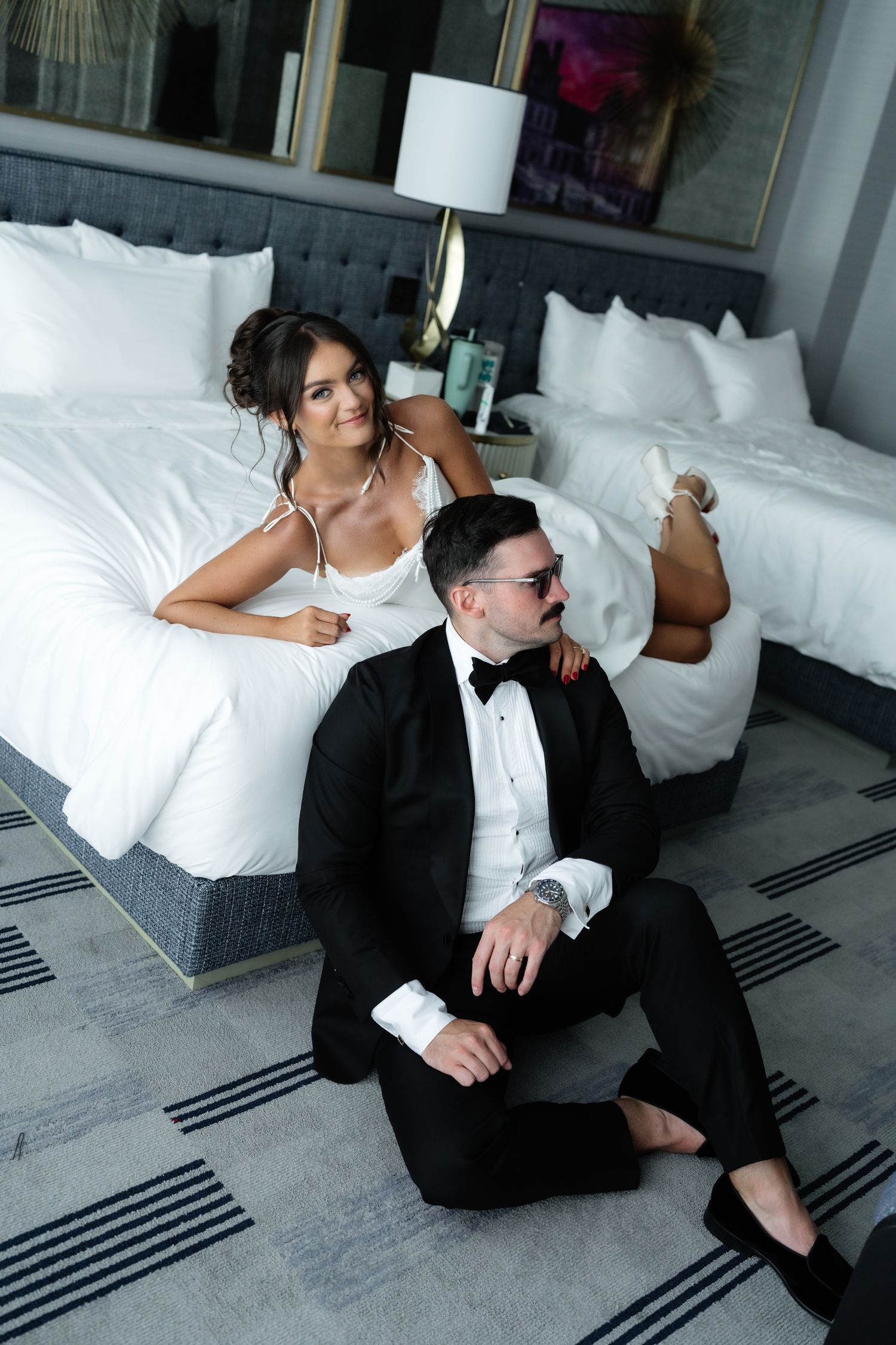 Bride in white gown reclines on hotel bed beside groom in black tuxedo sitting on floor.