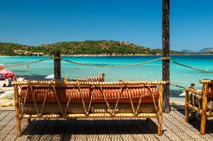 Bamboo daybed on a wooden deck overlooking a stunning turquoise beach with clear waters and green hills.