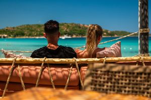 Couple relaxing in a hammock overlooking a turquoise tropical sea with lush green hills in the background.
