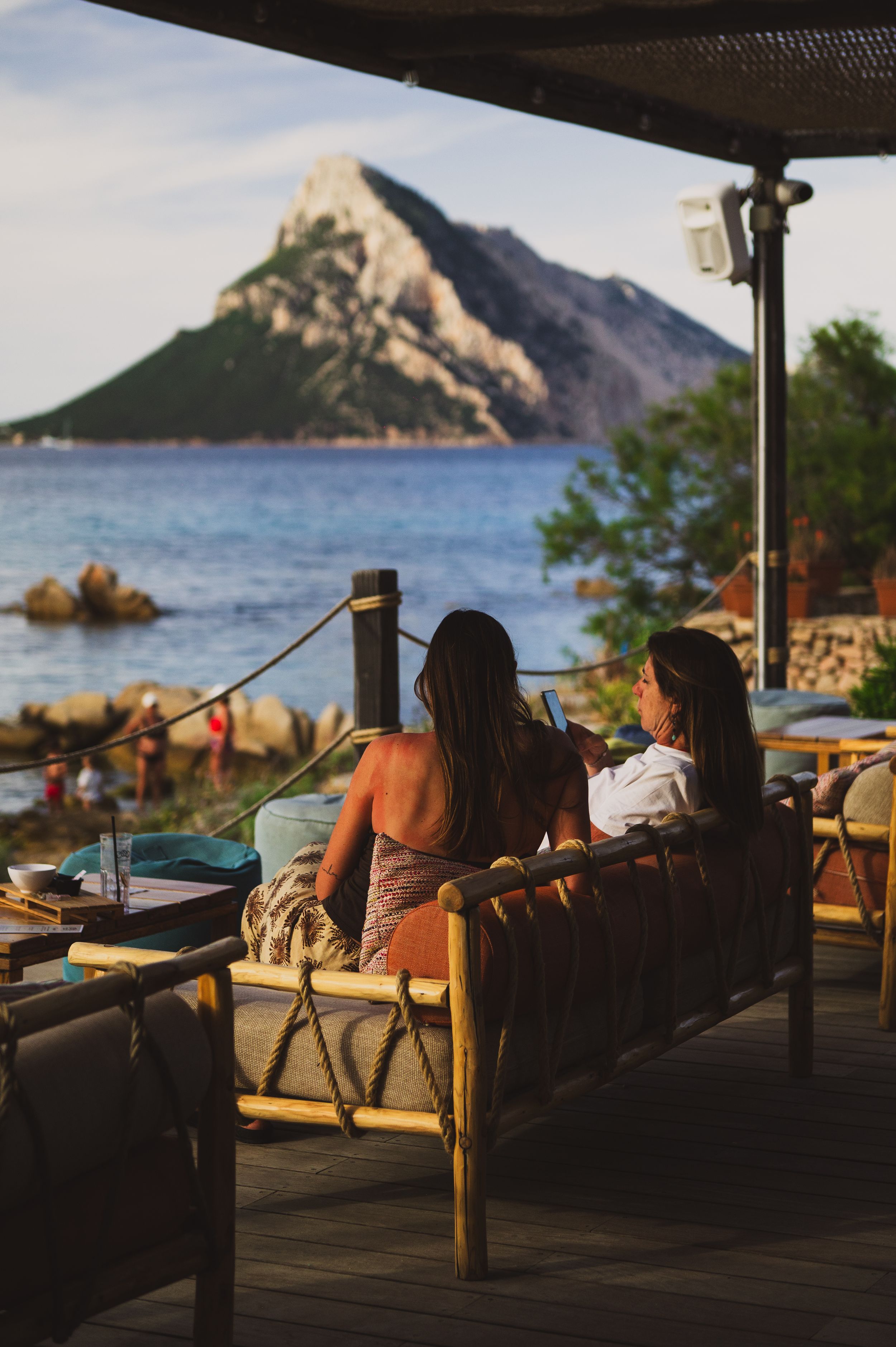 Two women sit on a bamboo sofa on a terrace overlooking a rocky island and blue sea at sunset.