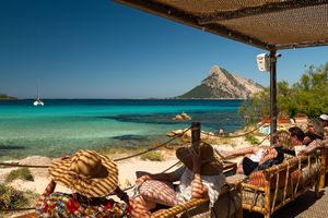 Tourists relaxing under a shaded terrace overlooking a turquoise Mediterranean bay with a sailboat and rocky island backdrop.