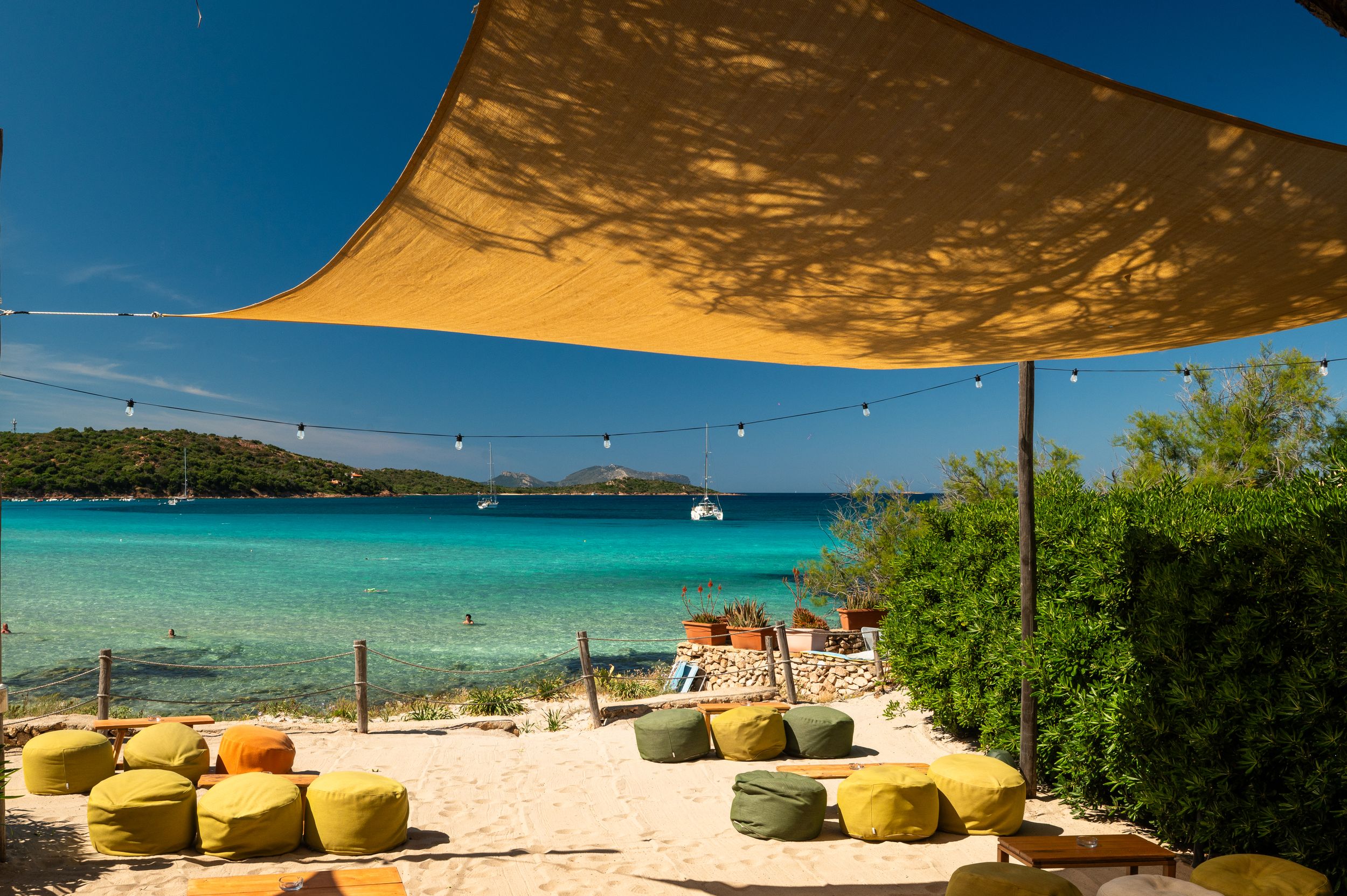 Tropical beach lounge with yellow shade sails, colorful poufs on white sand, and turquoise Caribbean waters under a clear blue sky.