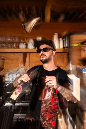 Tattooed bartender in sunglasses and black cap pours wine bottle with motion blur in a rustic bar setting.